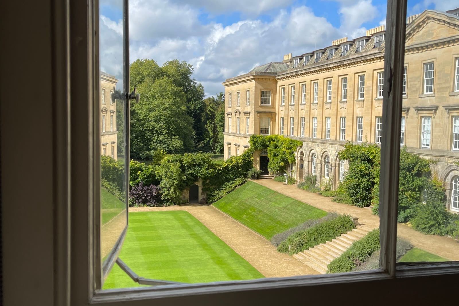 An open library window at Worcester College, Oxford, looking out over honey-colored stone buildings, a striped lawn, and a blue sky with clouds.
